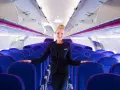 Uśmiechnięta stewardessa Wizz Air stoi w rzędach niebieskich foteli w samolocie.