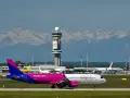 Samolot Wizz Air na płycie lotniska z wieżą kontroli lotów i górami w tle.