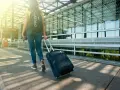 Kobieta z plecakiem i walizką na kółkach idzie w stronę nowoczesnego terminalu lotniska.