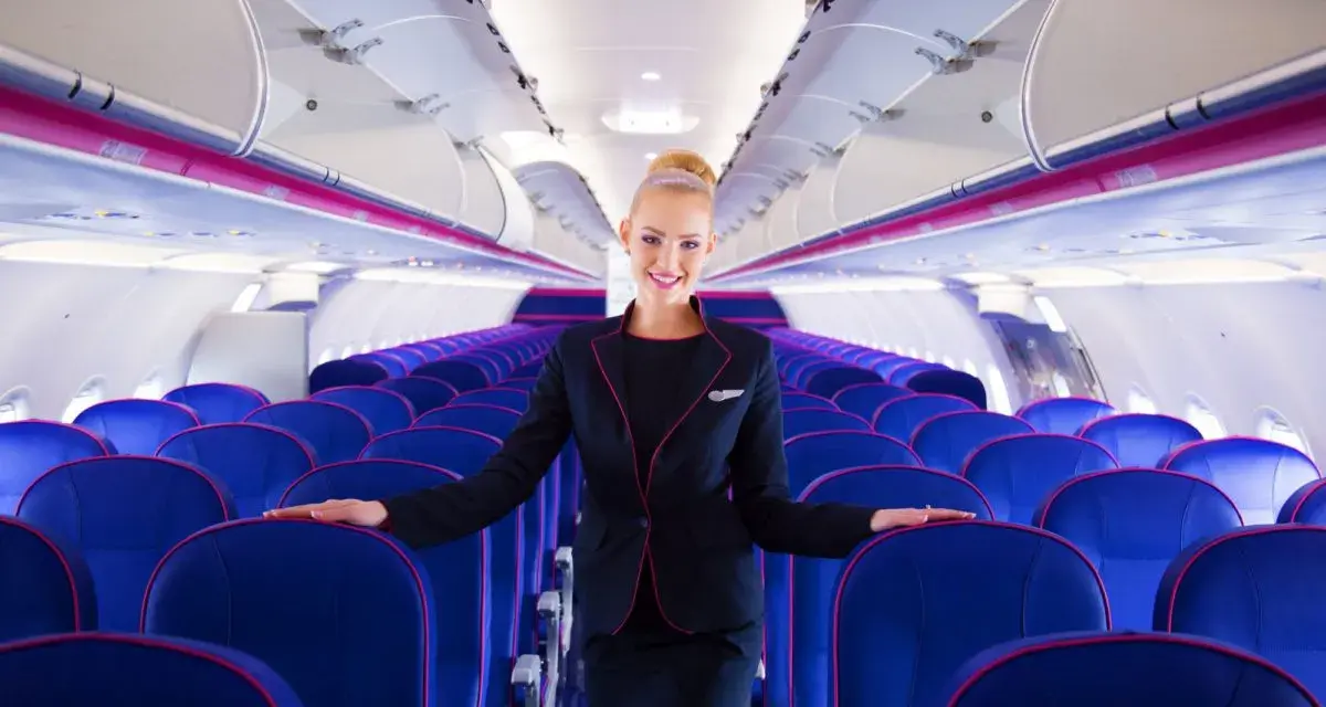 Uśmiechnięta stewardessa Wizz Air stoi w rzędach niebieskich foteli w samolocie.