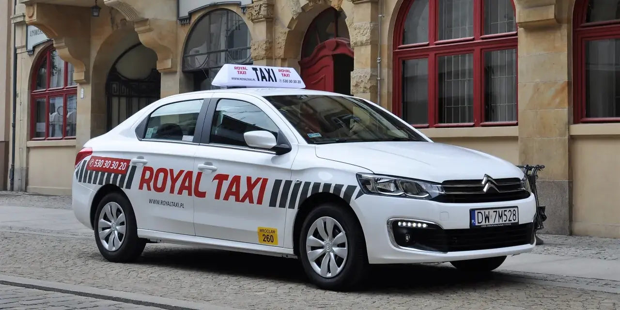 Biała taksówka Royal Taxi z numerem 260 na tle zabytkowej kamienicy we Wrocławiu.