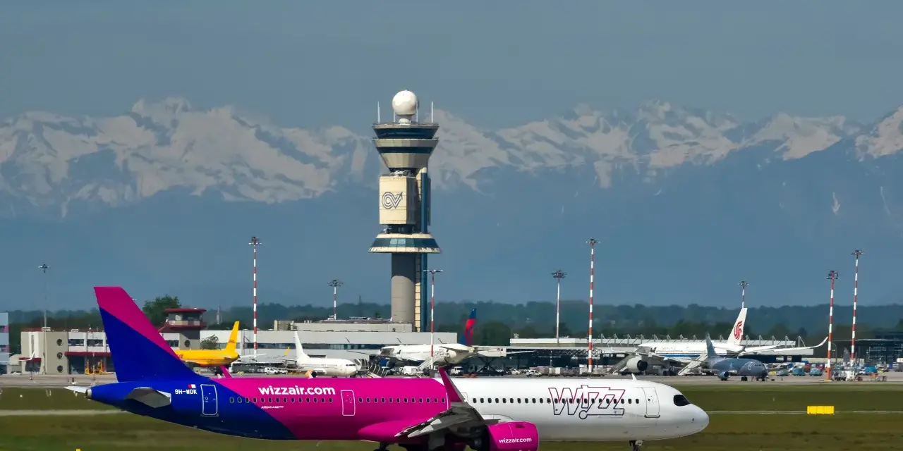 Samolot Wizz Air na płycie lotniska z wieżą kontroli lotów i górami w tle.