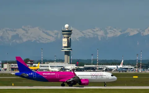 Samolot Wizz Air na płycie lotniska z wieżą kontroli lotów i górami w tle.