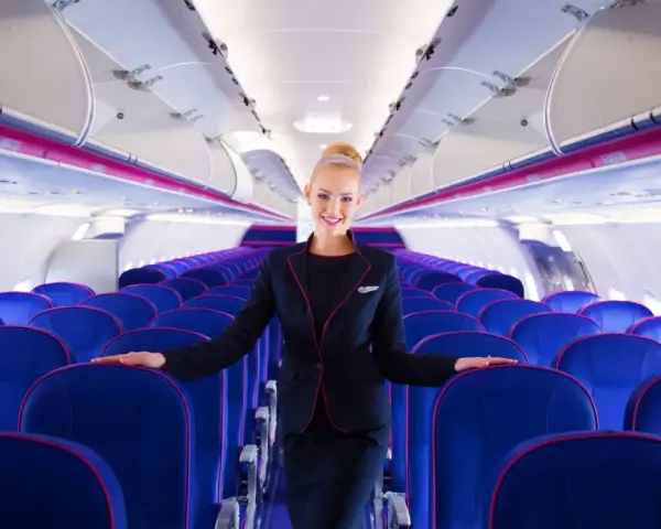 Uśmiechnięta stewardessa Wizz Air stoi w rzędach niebieskich foteli w samolocie.