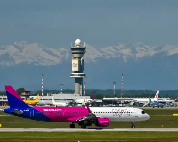 Samolot Wizz Air na płycie lotniska z wieżą kontroli lotów i górami w tle.