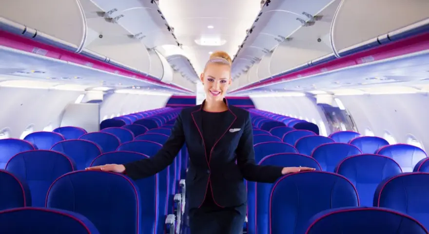 Uśmiechnięta stewardessa Wizz Air stoi w rzędach niebieskich foteli w samolocie.