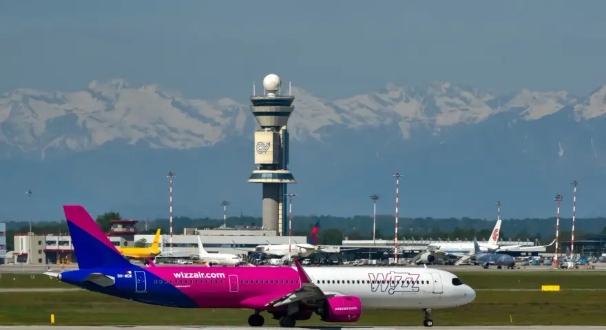 Samolot Wizz Air na płycie lotniska z wieżą kontroli lotów i górami w tle.
