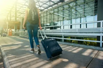 Kobieta z plecakiem i walizką na kółkach idzie w stronę nowoczesnego terminalu lotniska.